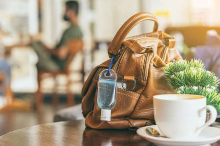 Mini alcohol gel bottle to kill Corona Virus(Covid-19) hang on a brown leather shoulder bag with hot coffee on table in cafe.New normal lifestyle.Health care concept.Selective focus on alcohol gelの写真素材