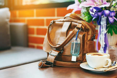 Mini alcohol gel bottle to kill Corona Virus(Covid-19) hang on a brown leather shoulder bag with hot coffee on table in cafe.New normal lifestyle.Health care concept.Selective focus on alcohol gelの写真素材