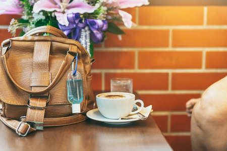 Mini alcohol gel bottle to kill Corona Virus(Covid-19) hang on a brown leather shoulder bag with hot coffee on table in cafe.New normal lifestyle.Health care concept.Selective focus on alcohol gelの写真素材