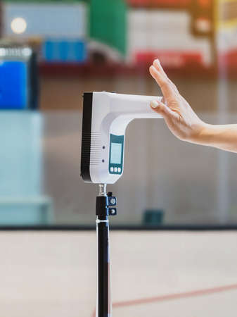 Customer with protective mask raised hand at digital thermometer with temperature scanning machine for fever before entering area. Screening to prevent infected people from entering restricted areas.の写真素材