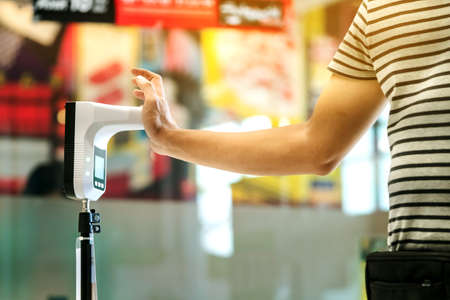 Customer with protective mask raised hand at digital thermometer with temperature scanning machine for fever before entering area. Screening to prevent infected people from entering restricted areas.の写真素材
