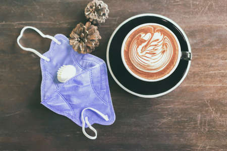 Hot Cappuccino coffee with protective mask on a wooden table for people being quarantined in the...