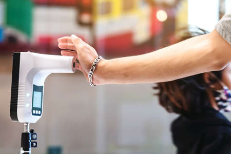 Customer with protective mask raised hand at digital thermometer with temperature scanning machine for fever before entering area. Screening to prevent infected people from entering restricted areas.の写真素材
