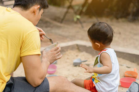Hand of father holds the food cup prepare to feed his toddler boy while playing sand together. Son and dad having happy nature moment time togetherness in summer. Father's day concept.のeditorial素材