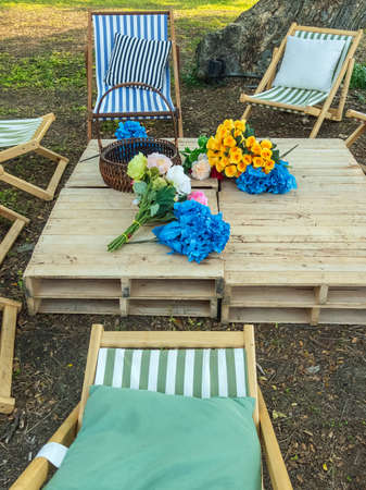 Beautiful artificial bouquets of various colors placed on wooden table among deck chairs in garden....