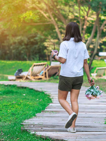 Back view of Asian woman with beverage and bouquet walk alone on pathway through green garden. Female relaxing alone in park. People spending time outside in green nature. Enjoying nature outdoors.の写真素材