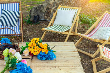 Beautiful artificial bouquets of various colors placed on wooden table among deck chairs in garden....