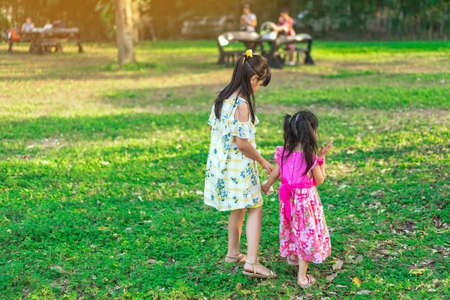 Back view of Asian little girls walking side by side through green garden.Elder sister and baby sister walking together in park. Happy family spending time together outside in green nature.の写真素材