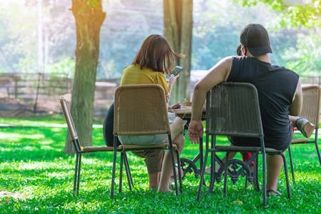 Back view of Asian young woman and her boyfriend sit to relaxing and chatting with friends in the garden. Happy friends spending time together outside in green nature. Enjoying nature outdoors.の写真素材