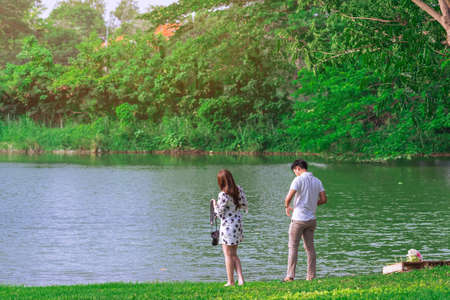 Back view of Asian lovers with camera relaxing near pond in garden. Summer vacation in green surroundings. Happy couple spending time together outside at waterfront in park. Enjoying nature outdoors.の写真素材