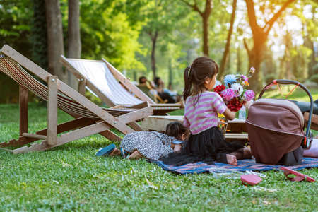 Back view of Asian young girls sit to relax together on mat near garden chair in garden. Summer vacation in green surroundings. Happy person outdoors relaxing on deck chair in garden. Outdoor leisure.の写真素材