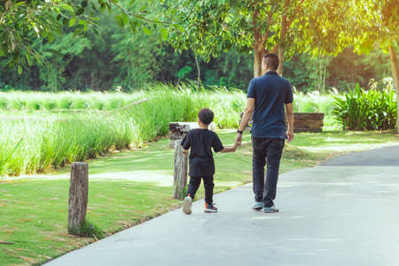Asian father hand holding lovely son walking on pathway through green garden.Dad and son walking together in park. Happy family spending time together outside in green nature.Enjoying nature outdoors.の写真素材