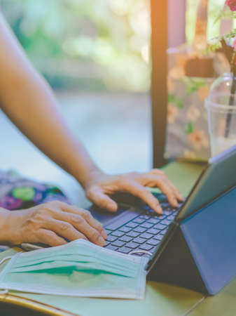 Hands of mature woman typing on laptop keyboard to work from home and take off her surgical mask put...