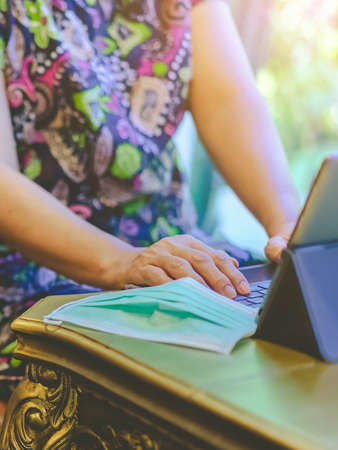 Hands of mature woman typing on laptop keyboard to work from home and take off her surgical mask put...