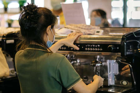 Back view of barista girl prepares coffee in cafe wearing surgical mask due to the pandemic of coronavirus (Covid-19). Young female barista in face mask and standing behind bar counter in coffee shop.の写真素材