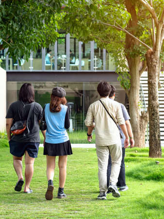 Back view of group of happiness Asian friends talking and walking along the path of park with green trees together. The friendship and fun of Asian friends spending their free time outdoors together.の写真素材