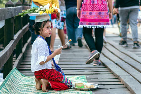 KANCHANABURI THAILAND-DECEMBER 31,2019 : Unidentified Mon girl eating sausage while sells flowers and make tanaka powdering service to tourists on Uttamanusorn wooden bridge at Sangkhlaburi.のeditorial素材