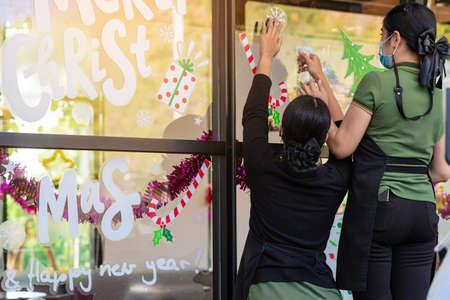 Asian women decorate beautiful stickers on window glass in front of cafe to welcome Christmas and New Year. Prepare to decorate the storefront to be beautiful during the important holidays of year.の写真素材