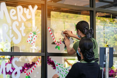 Asian women decorate beautiful stickers on window glass in front of cafe to welcome Christmas and New Year. Prepare to decorate the storefront to be beautiful during the important holidays of year.の写真素材