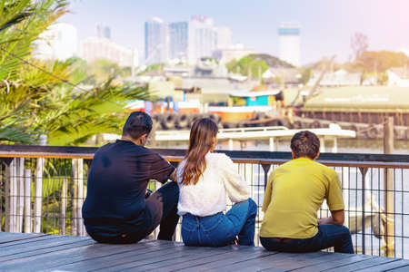 Back view of Asian friends relaxing and chilling at riverside. Relax lifestyle for friends enjoying freedom in summer holidays. Happy people outdoors resting at waterfront. Enjoying nature outdoors.の写真素材
