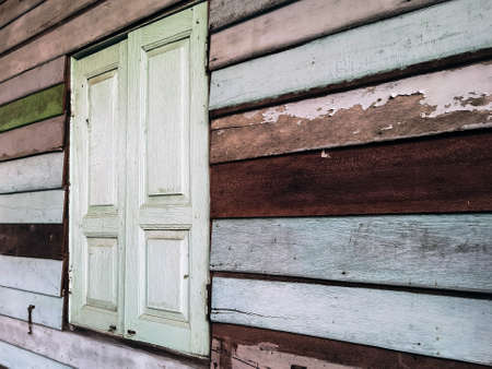 Old grunged wooden window frame painted white vintage with old colourful plywood wall. Antique window frame and old panes. Old closed window and planks of old wooden house. Background of wooden wallsの写真素材