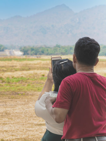Back view of happiness Asia couple tourists using  cell phone to take a selfie of themselves during an outdoor vacation in open field. Lovers take pictures together by using mobile phones in meadow.の写真素材