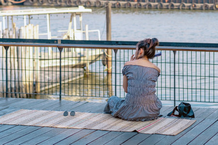 Back view of Asian woman relaxing and chilling at a riverside. Relaxed lifestyle for female enjoying freedom in summer holidays. Happy people outdoors relaxing at waterfront. Enjoying nature outdoors.の写真素材