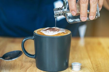 Close up of mature woman pouring warm syrup into hot cappuccino on cafe table. Pouring syrup in to...