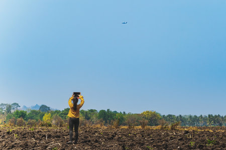 Back view of Asian young woman stand alone in the field holding up her arms with a tablet to take a photo and video clips an airplane is flying in the sky. Outdoor tablet photography technology.の写真素材