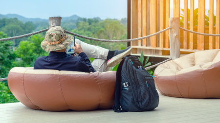 Male tourist wear hat with face mask with backpack use tablet while sit to relax on large cushions on balcony with blurred mountains in background. Traveller in bag chair enjoy tranquil outdoor natureの写真素材