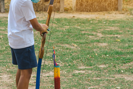 Back view of Asian boy wear face mask aims archery bow and arrow to colorful target in shooting range during training and competition. Exercise and concentration with outdoor archery. Archery for teenの写真素材