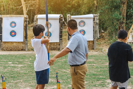 Back view of Asian boy wear face mask aims archery bow and arrow to colorful target in shooting...
