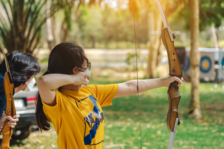Back view of Asian girl wear eyeglasses with face mask aims archery bow and arrow to colorful target in shooting range during training and competition. Exercise and concentration with outdoor archery.の写真素材