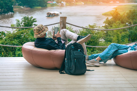 Male tourist wear hat with face mask and his wife use mobile phone and sit to relax on large cushions on balcony with mountains and river in background. Traveller in bag chair enjoy outdoor nature.の写真素材