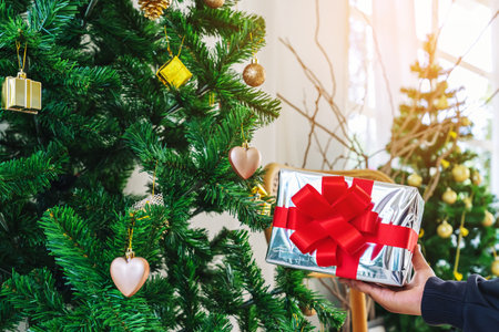 Beautiful craft paper gift box with vivid red ribbon bow over shiny sequins in young woman hands on background of beautifully decorated Christmas tree. Christmas and New Year concept. Selective focus.の写真素材
