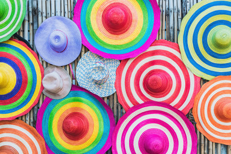 A variety of fashionable and colorful hats hanging on the bamboo backdrop of curbside shop. Women's designer hats from the sun of different colors. Design of women's beach hats. Beach hats for summer.の写真素材