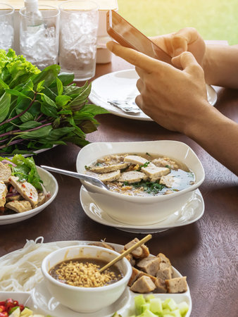 Hands of young man play a game with smart phone before eat pork sausage spicy salad Thai and Vietnam food (Yum Moo Yor) and noodle with other appetizing dishes and vegetables on wooden table.の写真素材