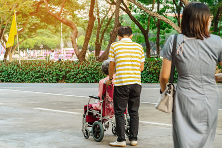 Back view of Asian elderly man walking with disabled elderly woman sitting in wheelchair outdoors wearing medical masks. Man pushes old lady in wheelchair through park. Healthy strong medical concept.の写真素材