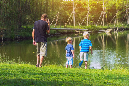 Back view of caucasian man use mobile phone to call about business and take care his son on vacation at waterside. Happy family outdoors resting together at waterfront. People enjoying nature outdoor.の写真素材
