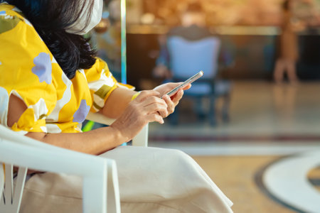 Hands of female traveler wear face mask sit to use smartphone while wait for hotel check in at hotel lobby. Business Woman waiting at hotel lobby using mobile phone. Business,travel and people conceptの写真素材