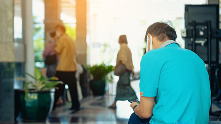 Back view of male traveler wear face mask sit to use smartphone while wait for hotel check in at hotel lobby. Businessman waiting at hotel lobby using mobile phone. Business, travel and people conceptの写真素材