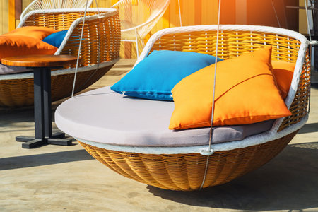 Colourful pillows on modern daybed. Brown rattan loungers with orange and blue pillows hanging with rope by relaxing terrace. Sunbed for sunbathing and swimming pool. Beautiful circular wooden daybed.の写真素材