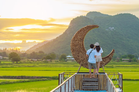 Happy couple take photo with crescent moon chair made of rattan for relaxation in paddy field with beautiful scenic in evening. Decorative wooden moon furniture as chair for viewpoint in rice field.の写真素材