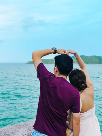 Happiness Asian lover with hands love sign on beach. Couple in love celebrates their engagement on seashore. Honeymoon trip. Lovers on beach. Wedding travel. Couple on vacation. Lover making heart.の写真素材