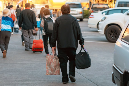 Back view of Asian elderly man traveler in black suit walks down a street with travel bag in morning. Senior male tourist walks with his travel bag. Vacation and travel elderly people concept. Journeyの写真素材