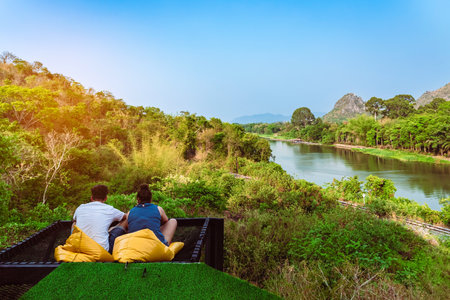 Back view of Happiness caucasian couple relaxing together on comfortable seats with beautiful view of nature at balcony on a hill near the riverbank. Happy love couple sitting together near riverside.の写真素材