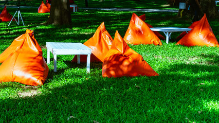 Beautiful view of orange triangle bean bags and cushions with white wooden table for customers to relax outdoors on green grass under shade of large tree with blurred background. Selective focus.の写真素材