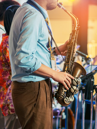 Musician playing saxophone on blurred background. Man with friends blow saxophone with the band for performance. Music instrument played by saxophonist player musician in festival. Selective focus.の写真素材