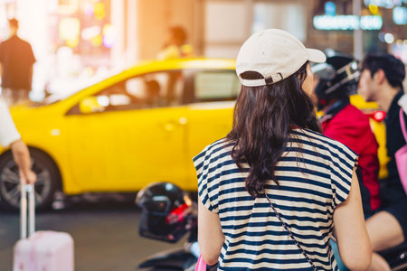 Back view of Asian woman wear white cap waiting taxi transports near city road with traffic on blurred urban background. Female traveller call taxi for tourism at airport. Traveling city alone conceptの写真素材