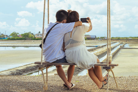 Asian couple sit on swing together and take a selfie while on vacation at salt field. Young lover making selfie photo with smart phone sitting together on swing with view of salt field in background.の写真素材